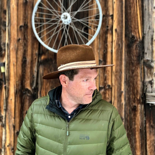 Adam Hevenor in a brown felt hat and olive down jacket, in front of a weathered barn wall