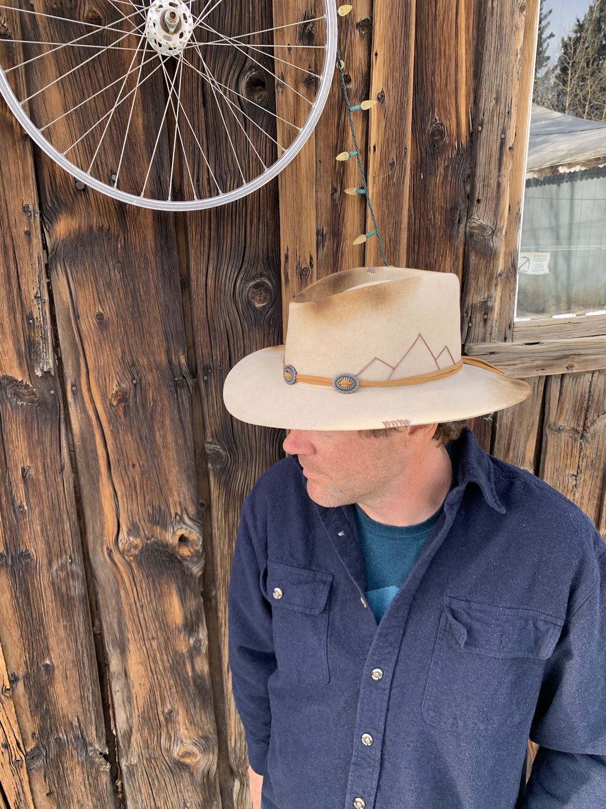 Adam Hevenor in a wide-brim felt hat and navy flannel, leaning against a weathered wood shed